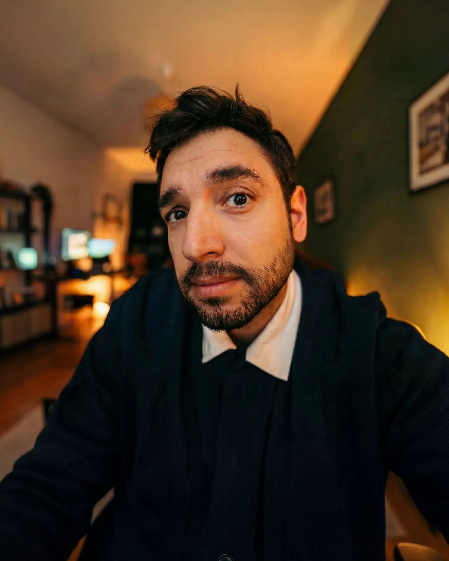 Erdem Güner im dunkelblauen Strickcardigan mit weißem Kragen, Selfie in seiner abendlich warm beleuchteten Wohnung